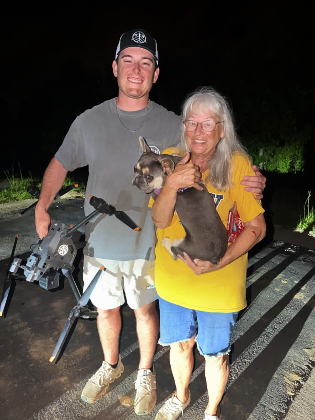 A woman holding her small dog Honey on a road at night after a thermal drone search, the drone on the ground beside her